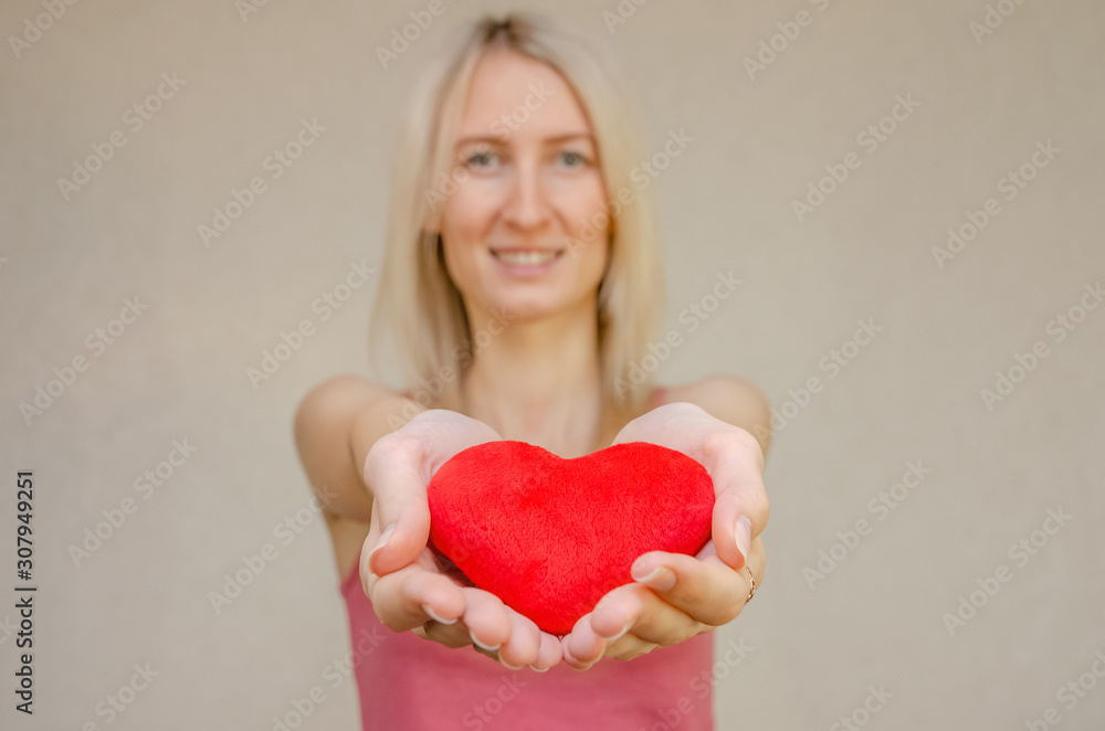 Blonde girl holding a red soft heart in front of her and smiling on a light background, copy space Blonde girl holding a red soft heart in front of her and smiling on a light background, copy space