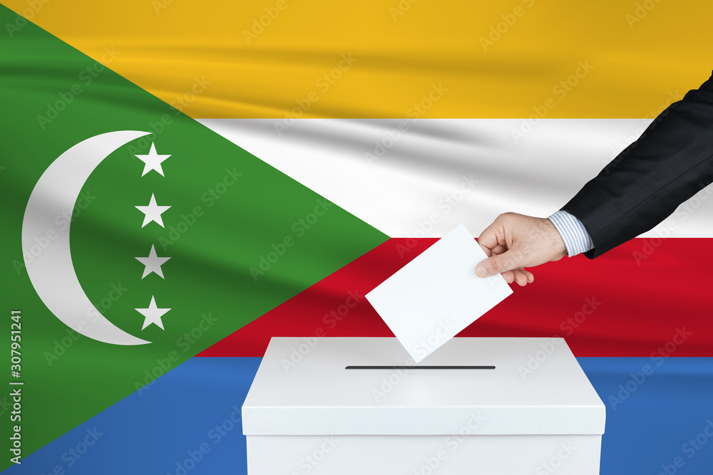 Election in Comoros. The hand of man putting his vote in the ballot box. Waved Comoros flag on background. Election in Comoros. The hand of man putting his vote in the ballot box. Waved Comoros flag on background.