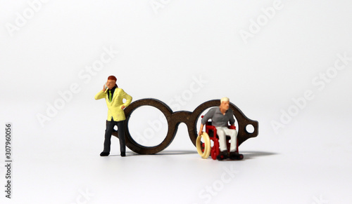 A miniature man in a wheelchair and a miniature man standing in front of a wooden miniature glasses.