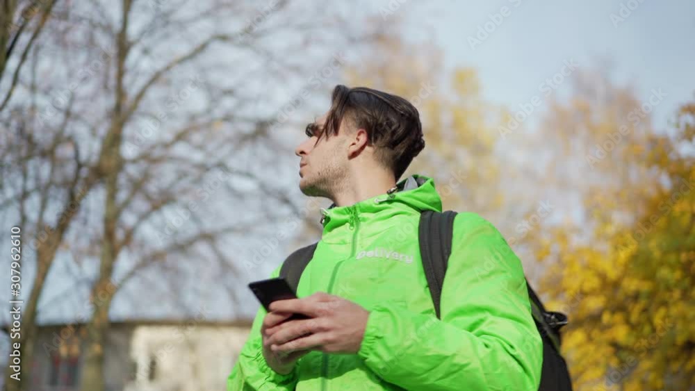 From below waist up shot of young food courier in green uniform using mobile navigation app on smartphone searching for direction standing in autumn street being lost From below waist up shot of young food courier in green uniform using mobile navigation app on smartphone searching for direction standing in autumn street being lost