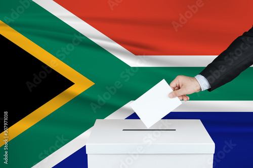 Election in South Africa. The hand of man putting his vote in the ballot box. Waved South Africa flag on background.
