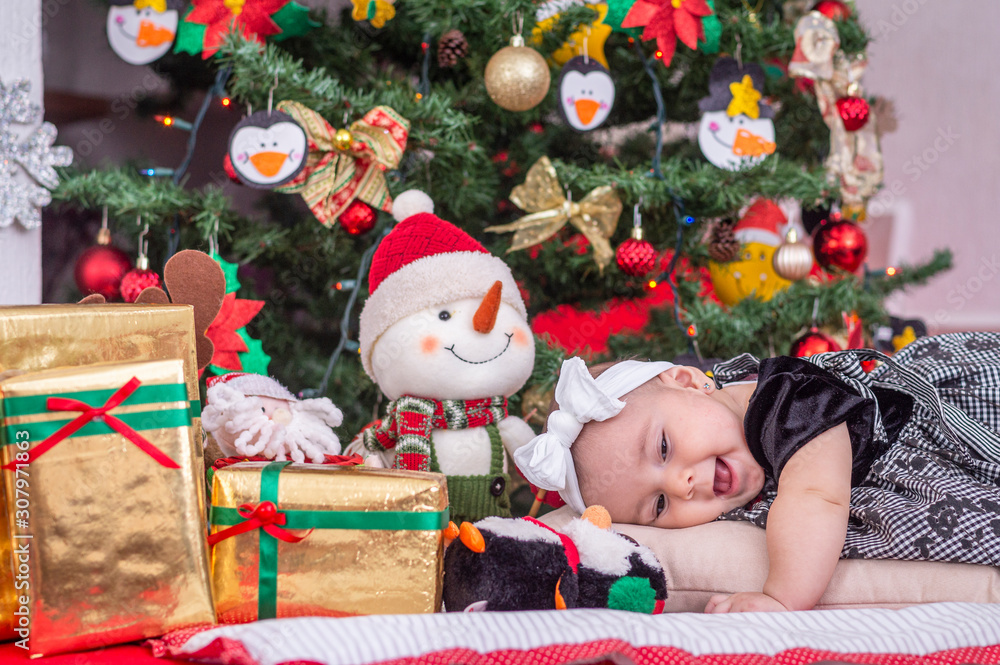 A white baby in Christmas tree decoration with black dress and white bow rests head on cushion and laughs A white baby in Christmas tree decoration with black dress and white bow rests head on cushion and laughs