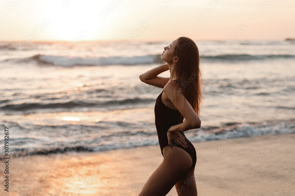 © Yevhenii - Backside view of girl with sexy booty in black bikini resting on deserted beach. Beautiful model in swimwear walks along  sandy beach on tropical island during sunset