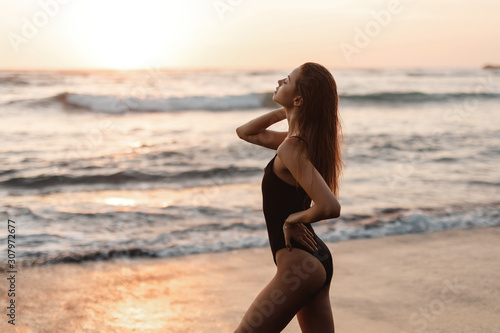 Backside view of girl with sexy booty in black bikini resting on deserted beach. Beautiful model in swimwear walks along sandy beach on tropical island during sunset