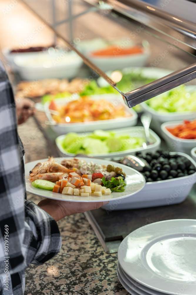 Vertical photo of a woman holding a plate of food and taking food with the other hand at a self-service restaurant