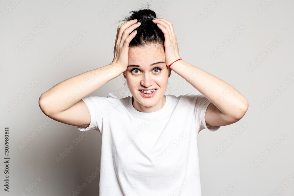 Shocked by an unexpected surprise, the woman smiles strangely and uncertainly, and looks at the camera, holding her hands on head. Mixed feelings and emotions. Shocked by an unexpected surprise, the woman smiles strangely and uncertainly, and looks at the camera, holding her hands on head. Mixed feelings and emotions.