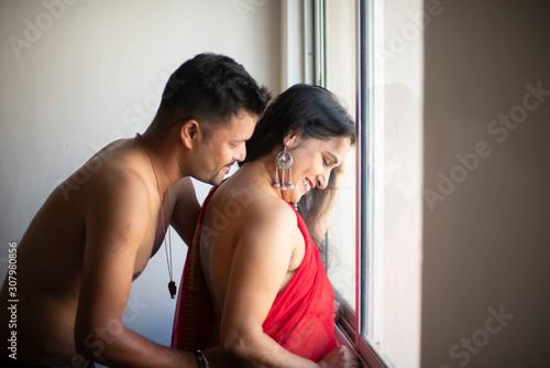 An young and handsome Indian brunette Bengali man trying to kiss his partner/girlfriend/wife from back in front of a glass window in white studio background. Indian lifestyle and bold fashion.