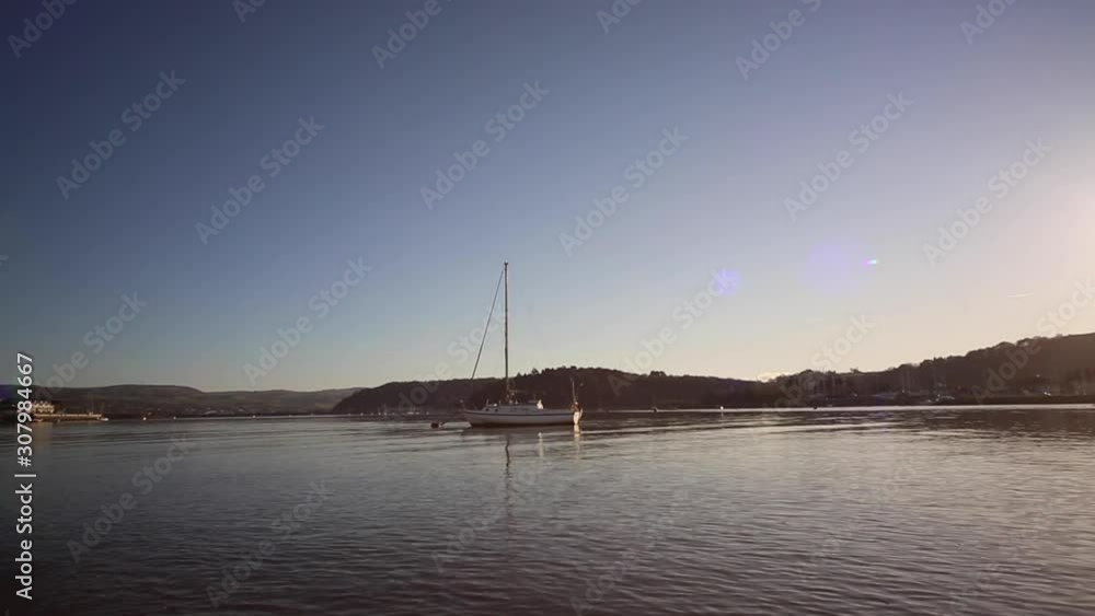 Distant view of a yacht bobbing up and down gently on its mooring buoy as the sun sets behind the mountains in the background Distant view of a yacht bobbing up and down gently on its mooring buoy as the sun sets behind the mountains in the background