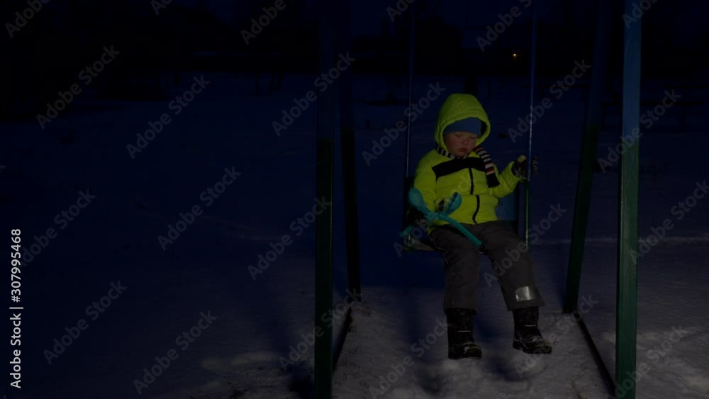 cute child in bright yellow jacket rides on swing on winter evening. Snowy winter in village. boy has fun alone in playground. Depressive evening lighting cute child in bright yellow jacket rides on swing on winter evening. Snowy winter in village. boy has fun alone in playground. Depressive evening lighting