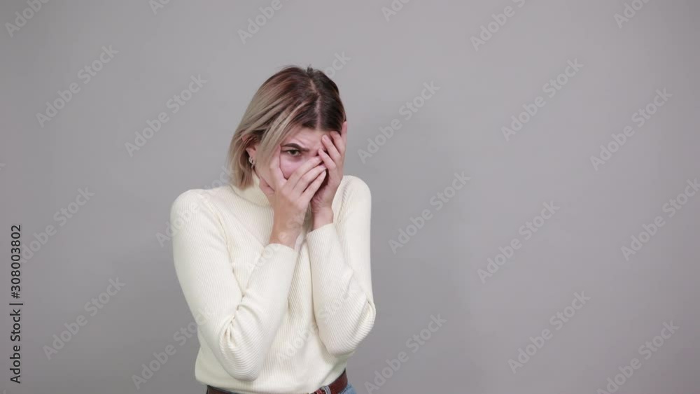 Bored young woman in casual clothes looking through fingers, with hand behind head isolated on gray wall background in studio. People sincere emotions, lifestyle concept. Bored young woman in casual clothes looking through fingers, with hand behind head isolated on gray wall background in studio. People sincere emotions, lifestyle concept.