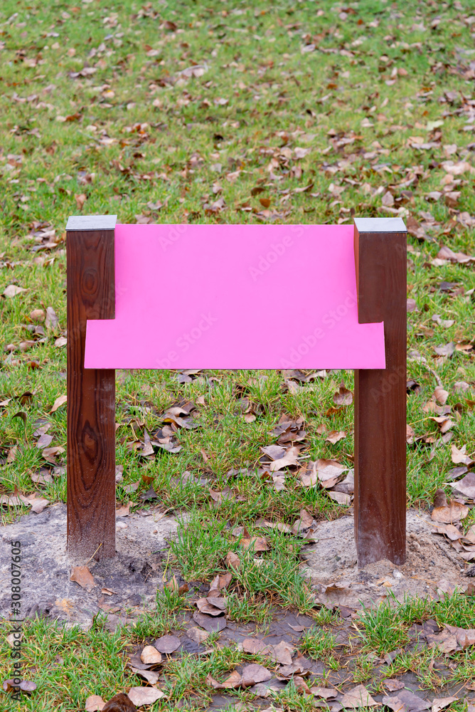 Blank pink wooden board for information and indications in the park, with a background of green grass covered by autumn leaves