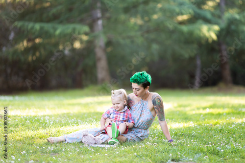 Young mother with green short hair is sitting on green grass, smiling, looking at her toddler son on her laps. Mother's day concept