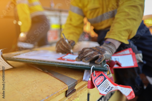 Personnel red danger locks attached with danger tags are locking on safety isolation permit lock box with defocused worker writing name, sign on prior locking on work permit safety control box