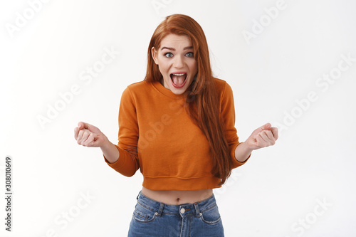 Surprised excited pretty ginger girl, female student winnin competition, achieve prize, clench fists and screaming from amazement and overjoy, winning, become champion, celebrating white background photo