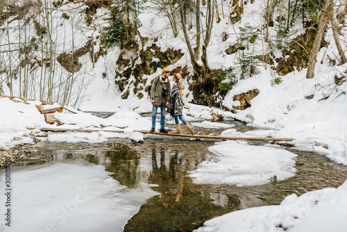 Man and woman happy couple hikers trekking in green winter forest and mountains. Young people walking on trail across the river , healthy lifestyle adventure, camping on hiking trip.