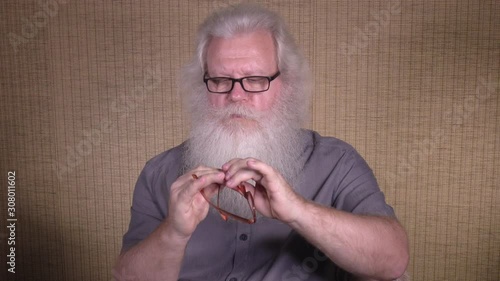Senior thickset Caucasian man with splendid grey hair and beard is repairing glasses with small screw. Domestic scene with elderly handsome European people, concentrated on homework, brown background.