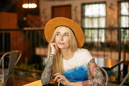 Photo of pretty young long haired woman with casual hairstyle sitting at table in cafe, looking aside positively while waiting for her order, wearing white and blue t-shirt and brown hat