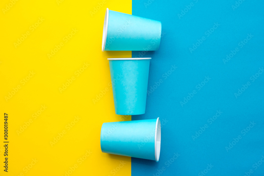 Obraz premium Blue paper disposable cups yellow and blue background. Cooking utensil. Cutlery sign. Top view. Minimalist Style. Copy, empty space for text