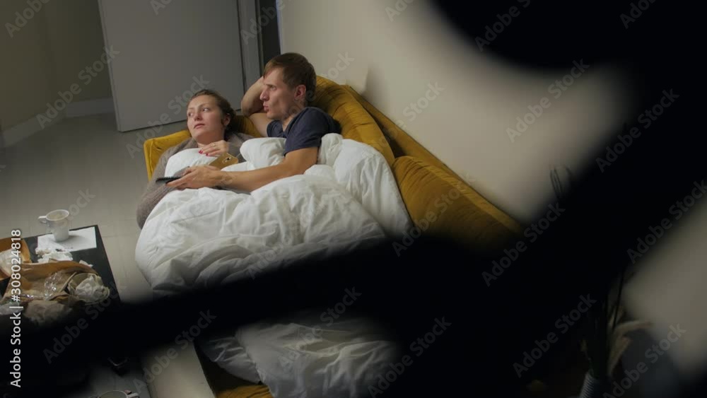 Young bored couple watching TV lay on yellow sofa woman and man covered white blanket fast food mess on table Young bored couple watching TV lay on yellow sofa woman and man covered white blanket fast food mess on table