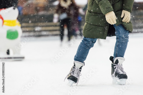 Male in olive jacket, blue jeans and in black sport skates riding on an outdoor ice rink. Snow is falling. Winter leisure and recreation. Selective focus, blurred background. Seasonal concept.
