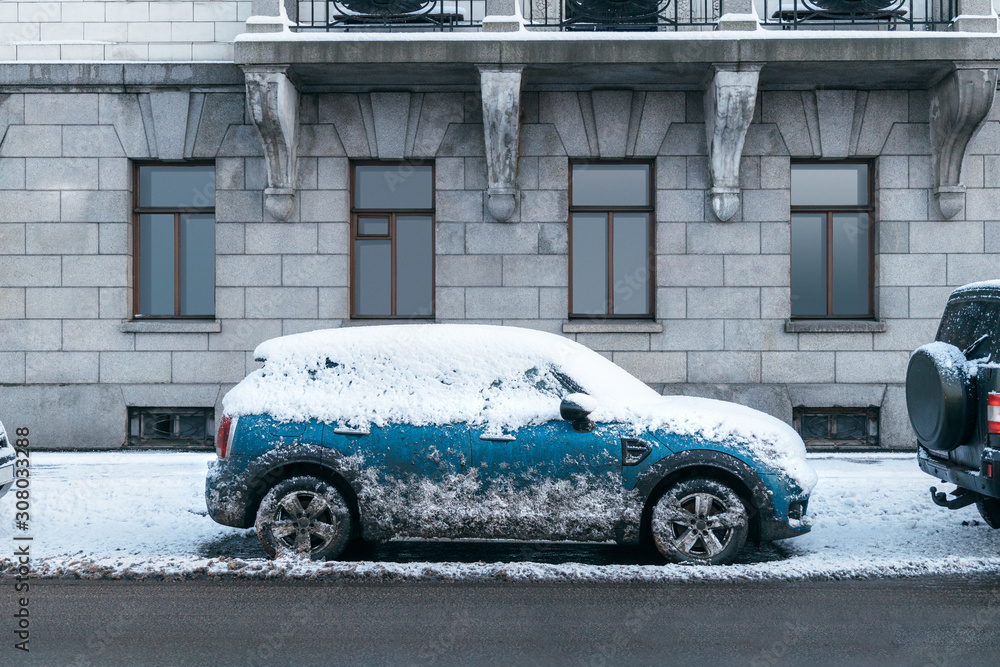 Photo of blue car covered with snow after heavy snowfall. Vehicle was not used for several days. Side of the car is stained with dirt because of passing cars. Winter and seasonal concept.
