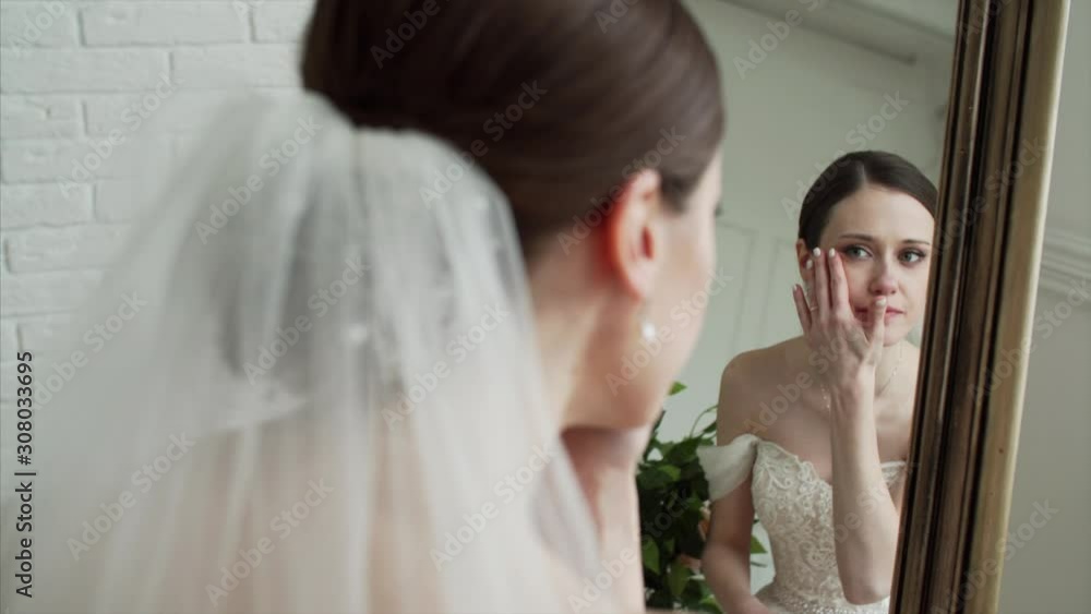 Young attractive bride in beautiful dress is looking into the mirror before the wedding ceremony Young attractive bride in beautiful dress is looking into the mirror before the wedding ceremony