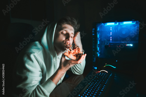 Young gamer sits at home at the table at the computer and eats pizza. Young man eating pizza at break after computer game. Night gaming with fast food photo