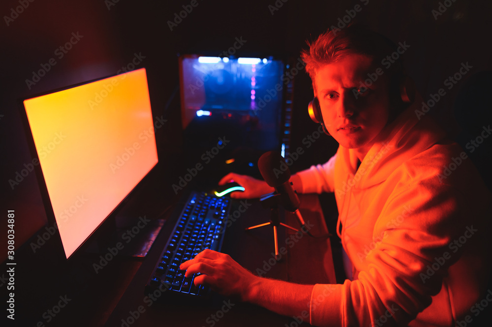Portrait of young gamer blogger in headphones with serious face sitting at  desk with microphone at computer with red light, looking into camera.  Streamer guy plays video games on his computer. Stock