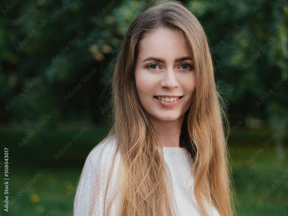 Close up headshot of adorable blonde haired charming smiling woman with blue eyes looking into camera in evening city park Close up headshot of adorable blonde haired charming smiling woman with blue eyes looking into camera in evening city park