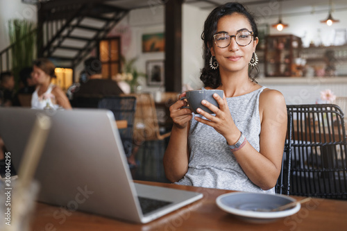 Relaxation, urban people and education concept. Charming young female freelancer, student having break from work, order coffee, holding cup and enjoy cappuccino, smiling pleased, using laptop