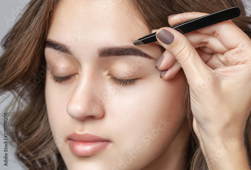 Make-up artist plucks eyebrows with tweezers to a woman with curly brown hair and nude make-up. Beautiful thick eyebrows close up. Professional makeup and cosmetology skin care. photo