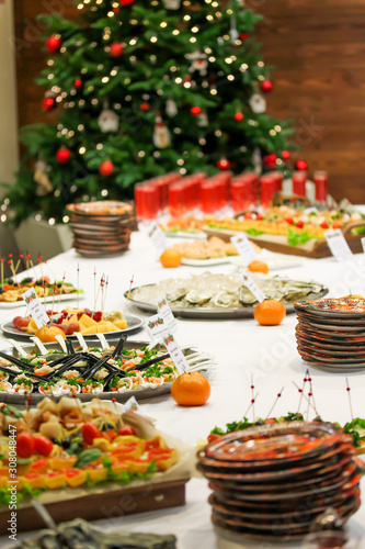 Christmas dishes and snacks at the party. Blurred decorated fir tree on the background. Bright festive food picture. Red drinks in glasses, sturgeon, red caviar, tangerines. Disposable plates