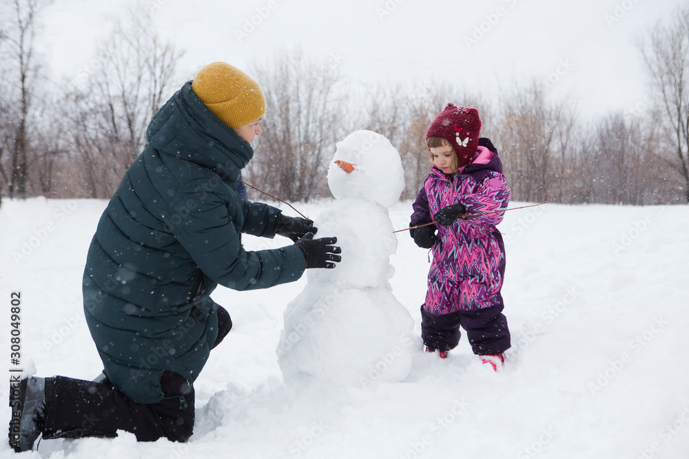 grandmother with the granddaughter make a snowman  together in winter park during snowfall