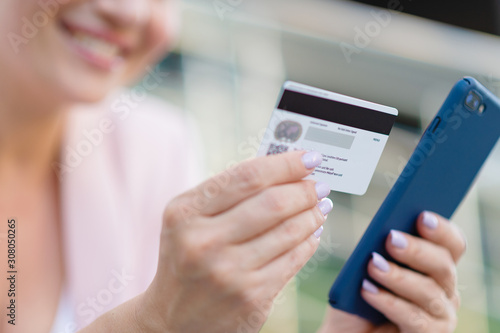 Businesswoman with a credit card and phone makes purchasing outdoors. Successful woman is using smart phone and bank card for online shopping. Beautiful smiling white woman holds card and cell phone
