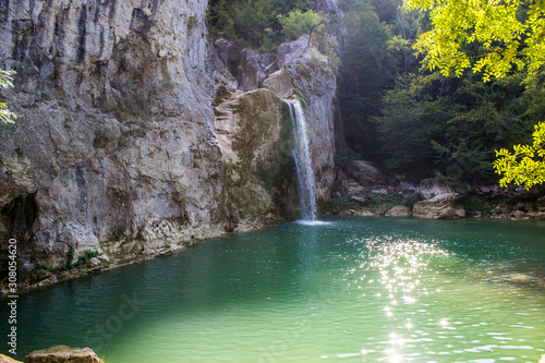 Little waterfall from rocky mountain falling to green lake in forest landscape. Summer time and sun flares on the lake.