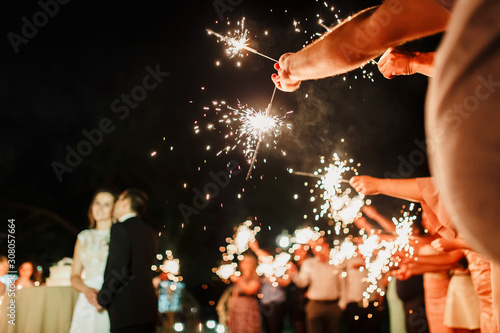 A crowd of young happy people with sparklers in their hands during celebration. Sparkler in hands on a wedding - bride, groom and guests holding lights in hand. Sparkling lights of bengal fires.