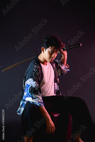 Asian man holding katana Japanese sword in his hand while sitting, ready to attack anytime. Eyes look serious on black background