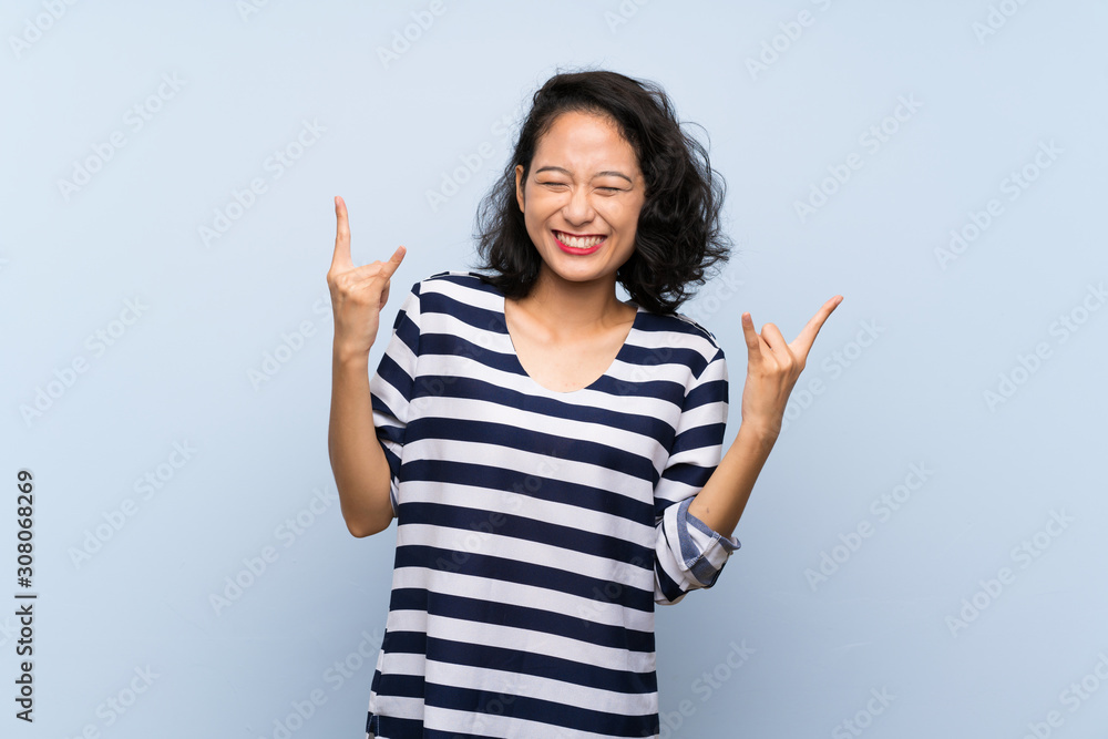 Asian young woman over isolated blue background making rock gesture Asian young woman over isolated blue background making rock gesture