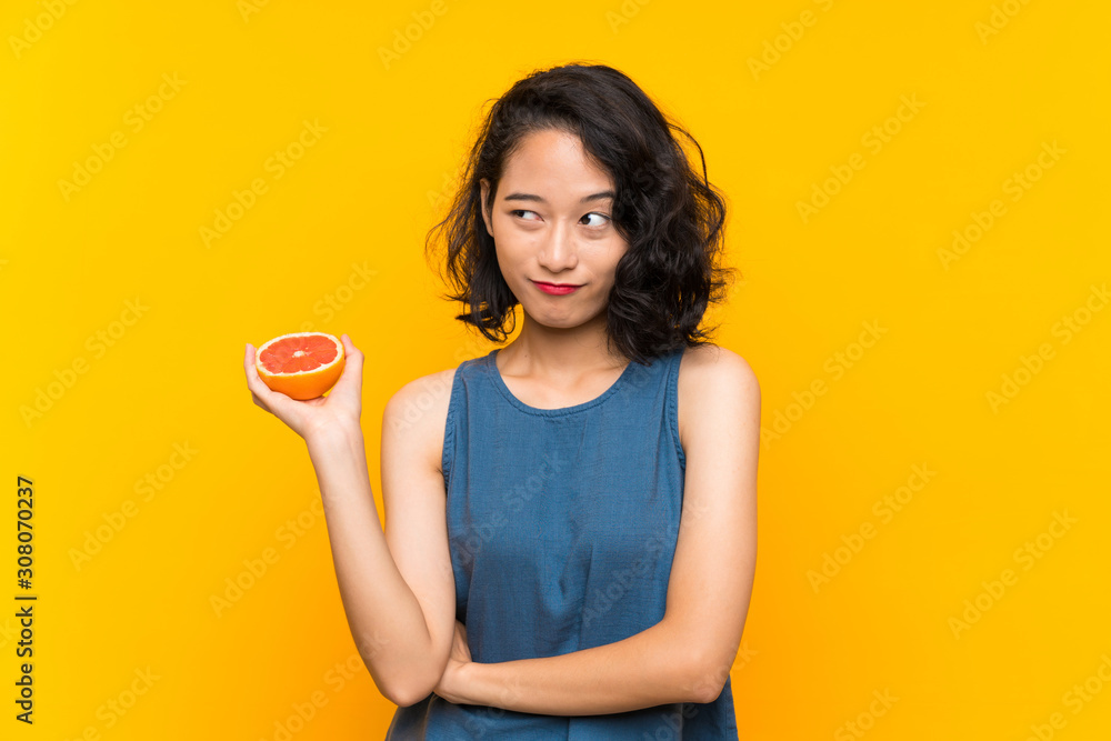 Young asian girl holding a grapefruit over isolated orange background making doubts gesture while lifting the shoulders