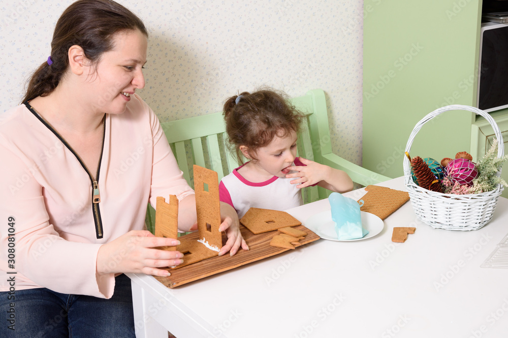 Mom glues the details gingerbread house with protein cream while her daughter looks. Mom glues the details gingerbread house with protein cream while her daughter looks.