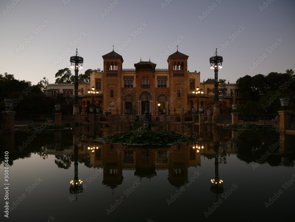 Reflection of a museum and lanterns in a pond during sunset, peaceful scene at Plaza America in Seville, Andalusia, Spain