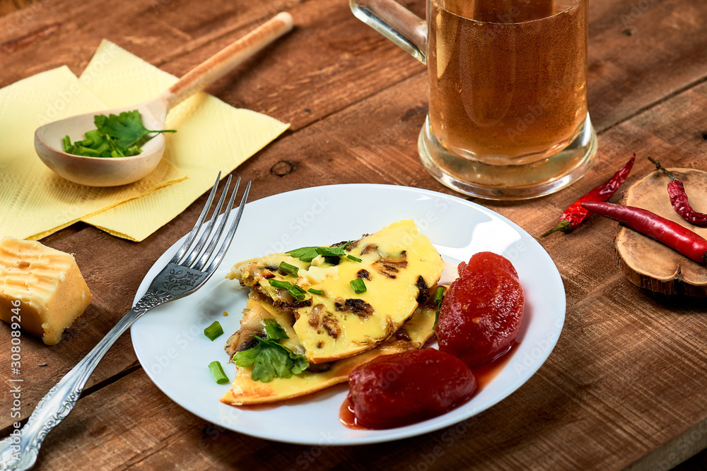 Scrambled eggs with cheese and mushrooms, green onions, parsley, tomatoes in a plate. Chili peppers and beer on a wooden table.