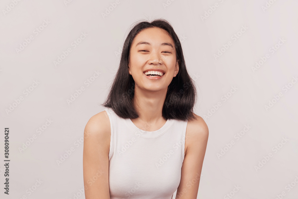 Girl laughs at something funny closes eyes with pleasure. Businesslike young woman Asian appearance with black hair and brown eyes dressed in short shirt stands isolated white background in Studio. Girl laughs at something funny closes eyes with pleasure. Businesslike young woman Asian appearance with black hair and brown eyes dressed in short shirt stands isolated white background in Studio.