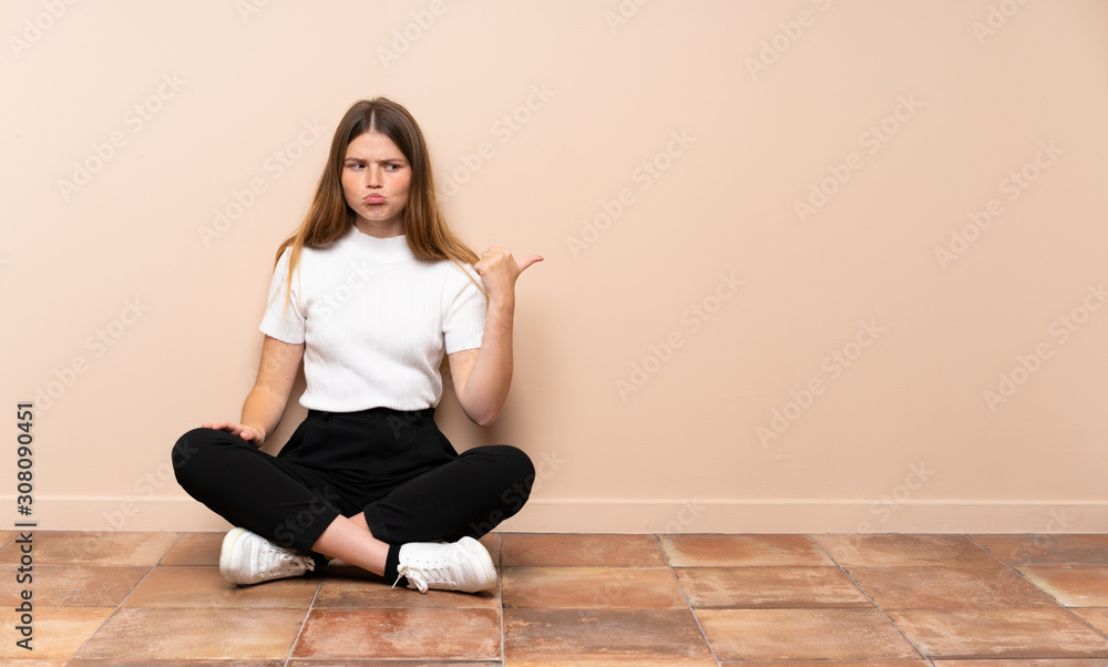 Ukrainian teenager girl sitting on the floor unhappy and pointing to the side Ukrainian teenager girl sitting on the floor unhappy and pointing to the side