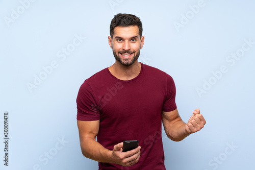 Handsome man over isolated blue background with phone in victory position