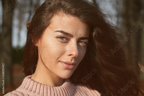 Warm autumn. Portrait of young woman with brown hair, green eyes wearing pink sweater walking in colorful forest or Park. Girl looking into camera has natural beauty maple leaves in hair develops wind