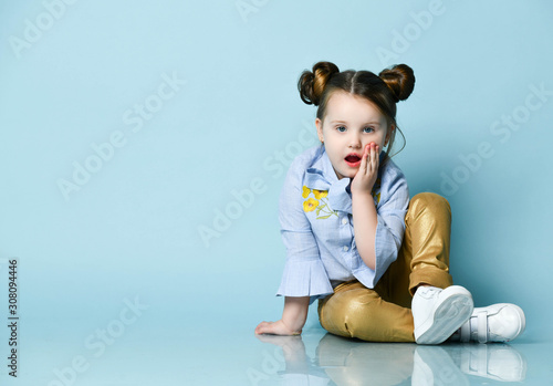 Surprised little baby kid girl in blue shirt and gold leather pants is sitting on the floor holding hand at her cheek