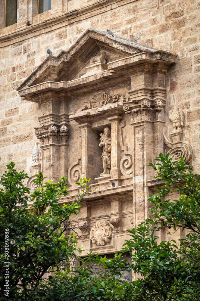 Typical historical facade of Valencia old town with beautiful statue on the background and green trees at the front.