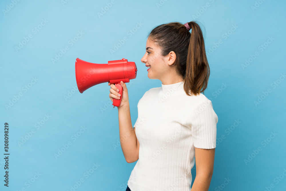 Young brunette girl over isolated blue background shouting through a megaphone Young brunette girl over isolated blue background shouting through a megaphone