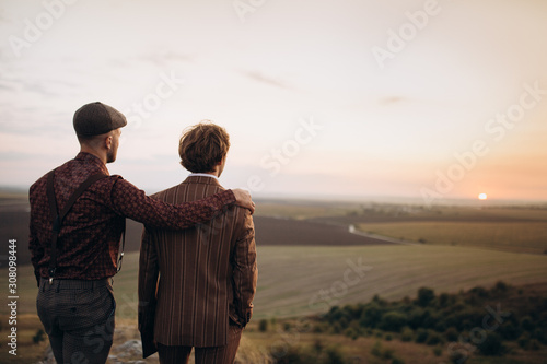Two guys in suits standing on a hill looking towards a deep orange sky and beatiful sunset. Rear view, hand on friend's shoulders. Concept of support and friendship. Copy space